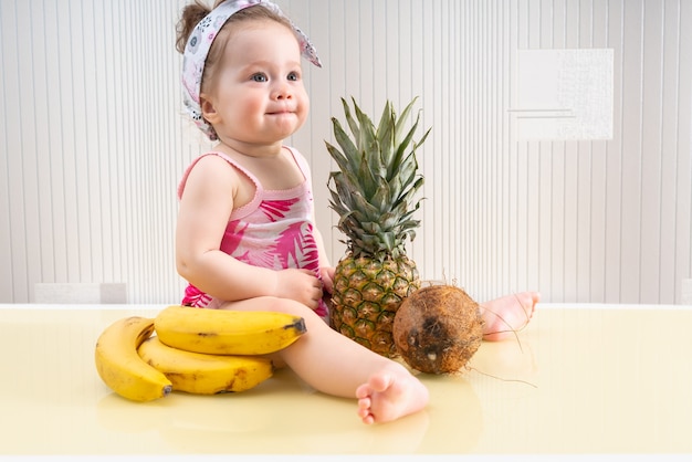 Cute little baby in pink shirt sitting among tropical fruits