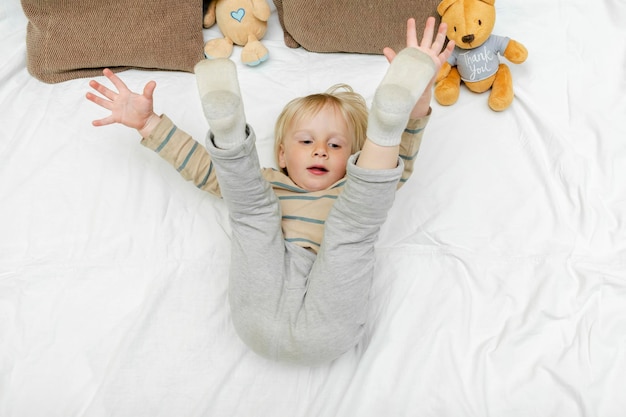 Cute little blond caucasian child kid toddler boy playinghaving fun lying on bedlifts up the legs at home at sunny morning wakes up in his room happy childhood moments