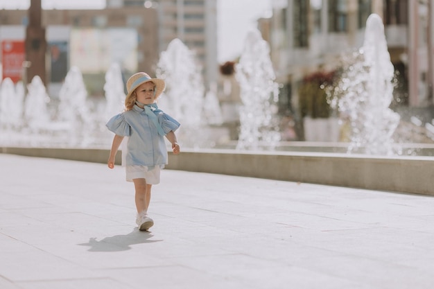 cute little blond girl in blue dress and straw hat playing near fountain with plush bunny, card