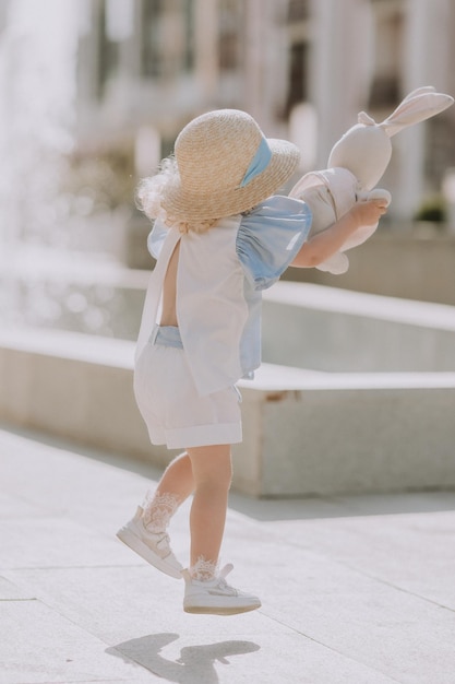 cute little blond girl in blue dress and straw hat playing near fountain with plush bunny, card