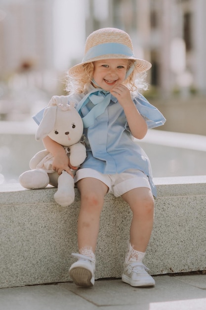 cute little blond girl in a straw hat and a blue dress plays with a stuffed rabbit outdoors