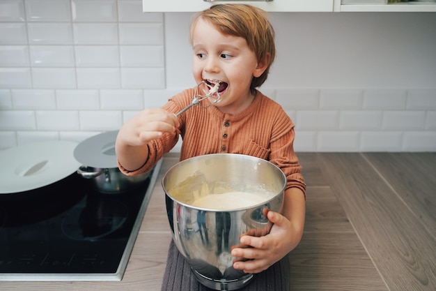 Cute little blond preschool kid boy baking cake in domestic kitchen indoors Funny child tasting sweet dough Little helper