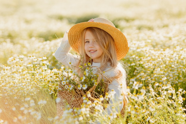 Cute little blonde girl in a cotton dress and straw hat walks in a field of daisies collects them in the basket