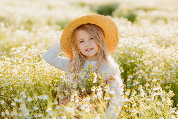 Cute little blonde girl in a cotton dress and straw hat walks in a field of daisies collects them in the basket