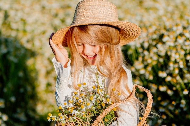 Cute little blonde girl in a cotton dress and straw hat walks in a field of daisies collects them in the basket