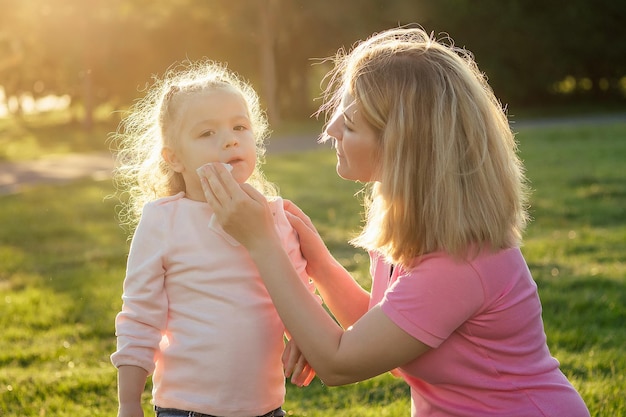 Cute little blonde girl daughter in a blue dress with beautiful mother applying antiseptic antibacterial gel in summer day in field greens grass background protection from germs at picnic