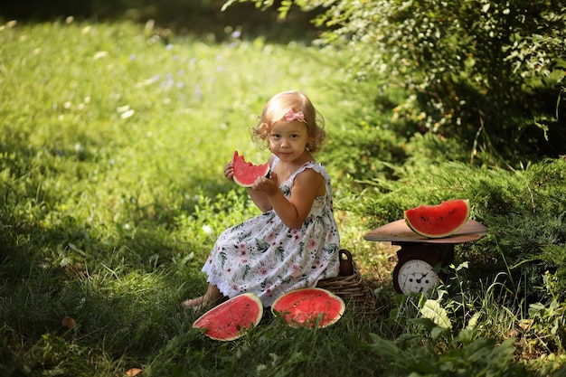 A cute little blonde girl eats a fresh red watermelon in the summer on the street