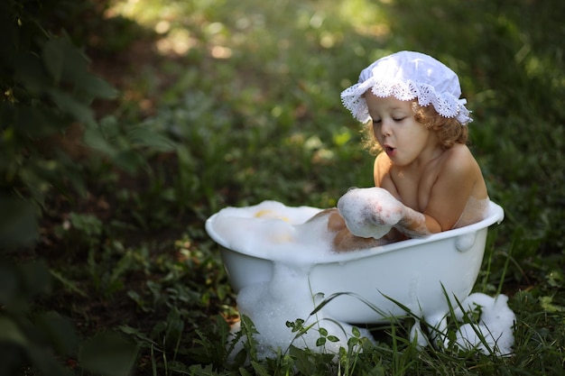 A cute little blonde girl is sitting in the bathroom with foam and ducklings in the summer on the s