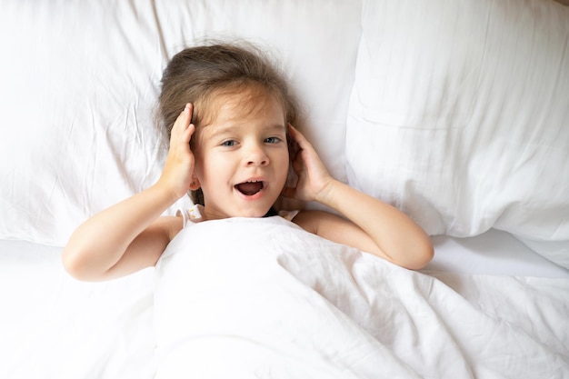 A cute little blueeyed girl yawns in the morning in a white bed A sleepy child Mode of the day Lifestyle Space for text