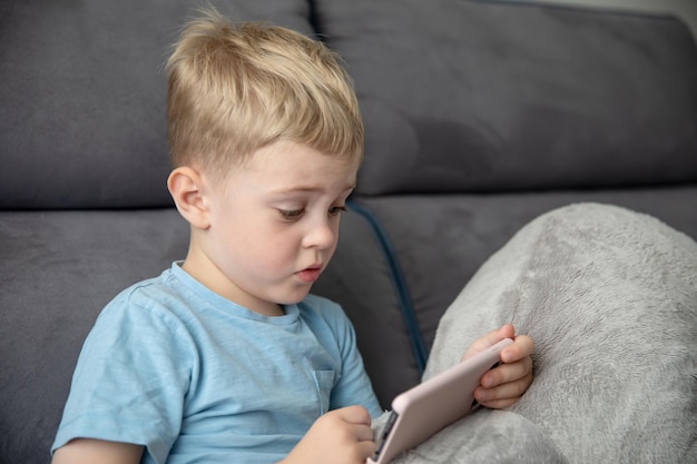 Cute little boy blonde looks at the smartphone sitting on the couch at home. Concept online education, learning, games