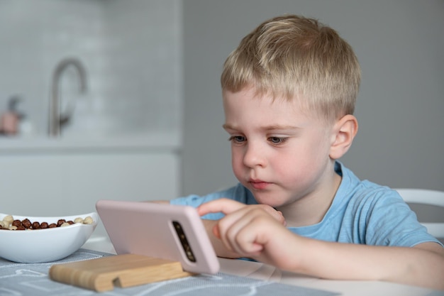 Cute little boy blonde looks at the smartphone sitting at the kitchen at home. Eats breakfast cereal. Concept online education, learning, games.