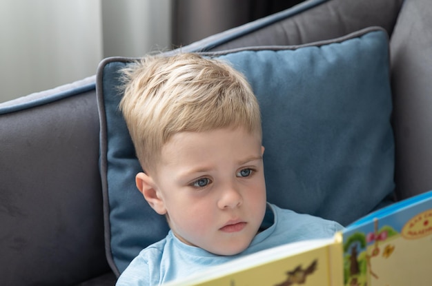 Cute little boy blonde reading a book lying on the couch