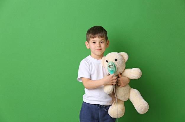 Cute little boy holding nebulizer and toy bear on color background Allergy concept