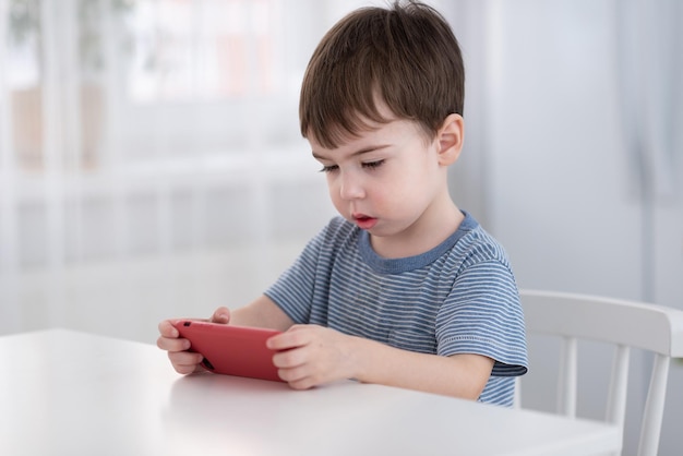 A cute little boy is sitting at the table and playing on the phone Gadgets and kids concept