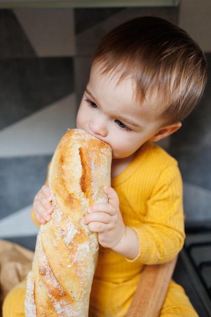 A cute little boy one year old sits and eats freshly baked rye bread The child holds a fresh baguette in his hands