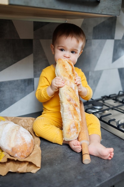 A cute little boy one year old sits and eats freshly baked rye bread The child holds a fresh baguette in his hands