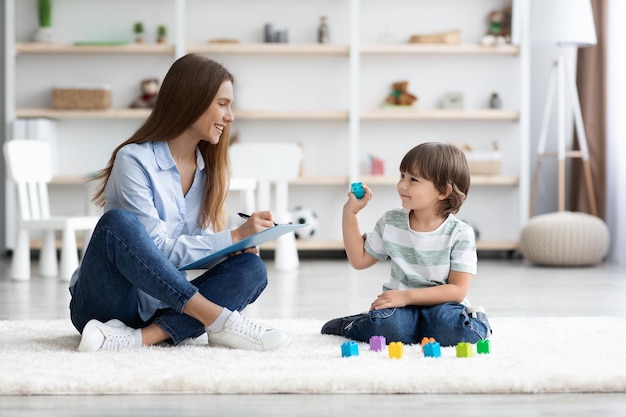 Cute little boy playing with colorful blocks young professional woman psychologist making notes