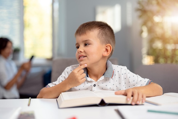 Cute little boy sitting at desk with books and notes and dreaming about something Caucasian schoolboy looking aside during domestic education