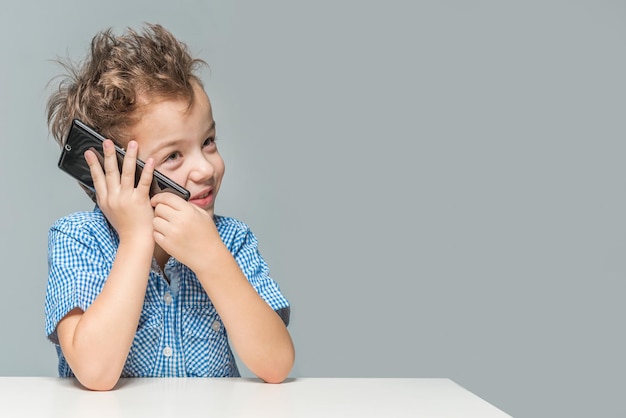 Cute little boy talking on the phone while sitting at the table on a gray background Isolated