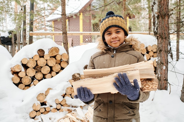 Cute little boy in warm winter jacket and knitted beanie holding firewood