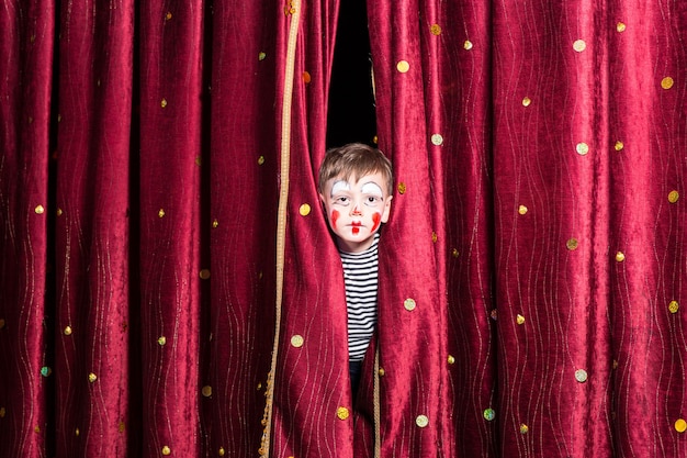 Cute little boy wearing colorful red face paint and a costume peering out from between the curtains on stage as he waits for a pantomime to begin