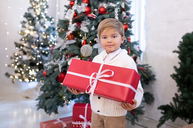 Cute little boy with Christmas present gift box near Christmas trees with lights