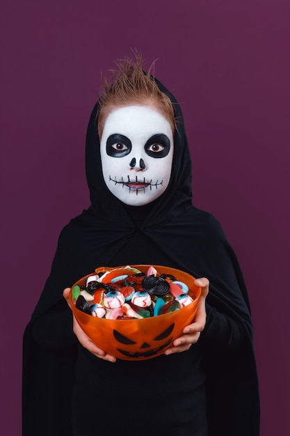 Cute little boy with halloween makeup dressed as a skeleton holding a cup of sweets.