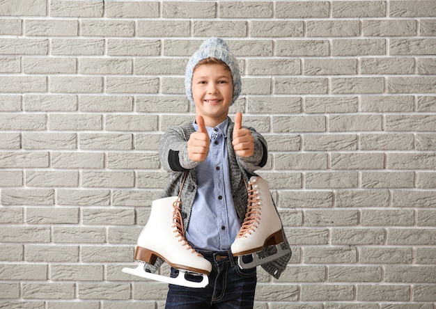 Cute little boy with ice skates showing thumb-up against brick