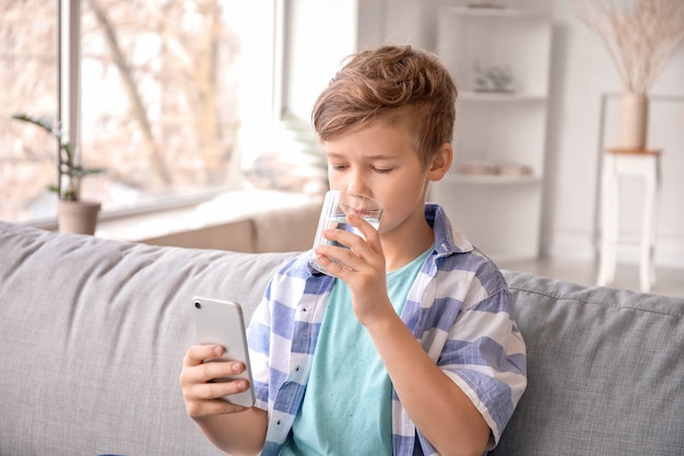 Cute little boy with mobile phone drinking water at home