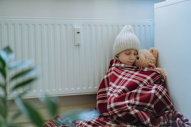 Cute little boy wrapped id plaid wearing knit hat sitting by heater hugging teddy bear cold at home