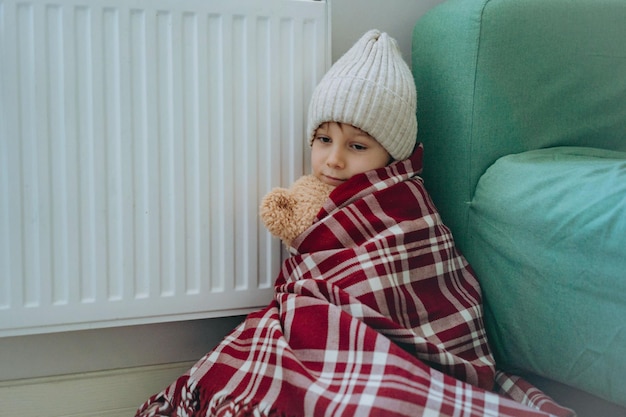 Cute little boy wrapped id plaid wearing knit hat sitting by heater hugging teddy bear cold at home