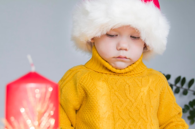 A cute little boy in a yellow sweater and a red Santa Claus hat with a red Christmas candle