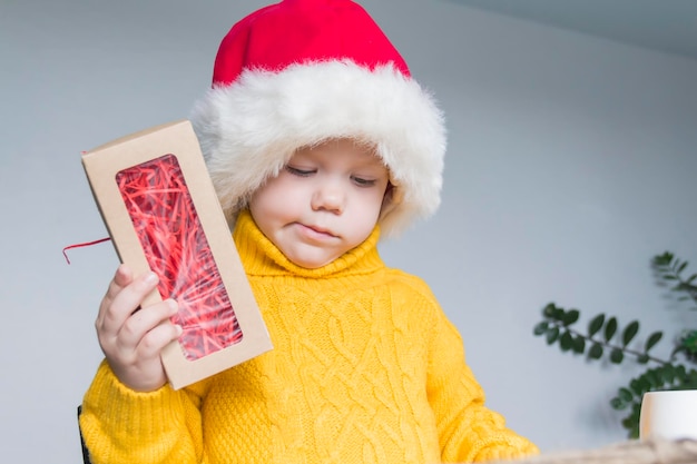 A cute little boy in a yellow sweater and a red santa claus hat at a wooden table with gifts that ar