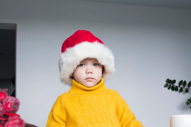 A cute little boy in a yellow sweater and a red Santa Claus hat at a wooden table with gifts that are packed in kraft paper