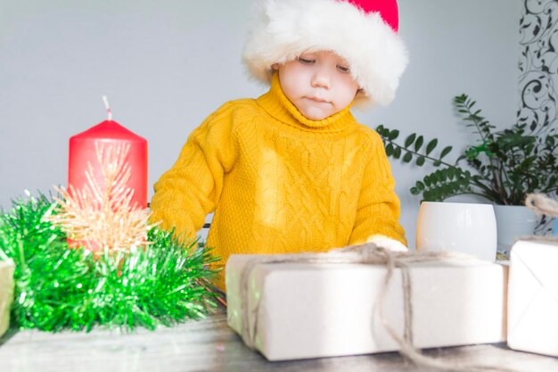 A cute little boy in a yellow sweater and a red Santa Claus hat at a wooden table with gifts that are packed in kraft paper