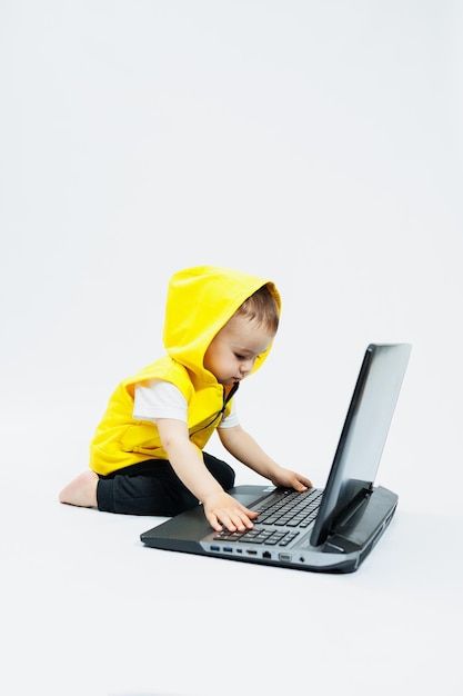 A cute little boy in a yellow vest is sitting at a digital laptop on a white background Child and modern portable computer gadget