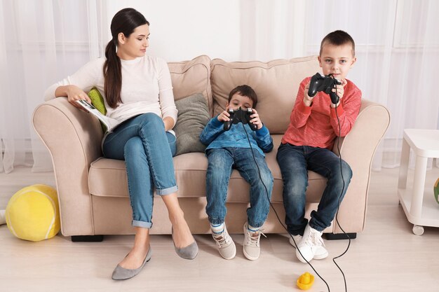 Cute little boys playing video game while their nanny is reading magazine at home