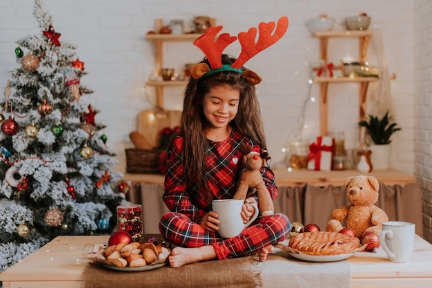 Cute little brunette girl in red pajamas with reindeer horns on her head is eating a Christmas cake