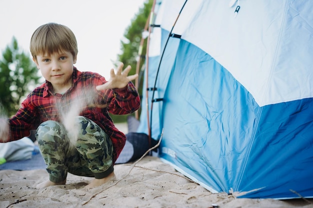 Cute little caucasian boy sitting on sand barefoot by touristic tent Family camping concept