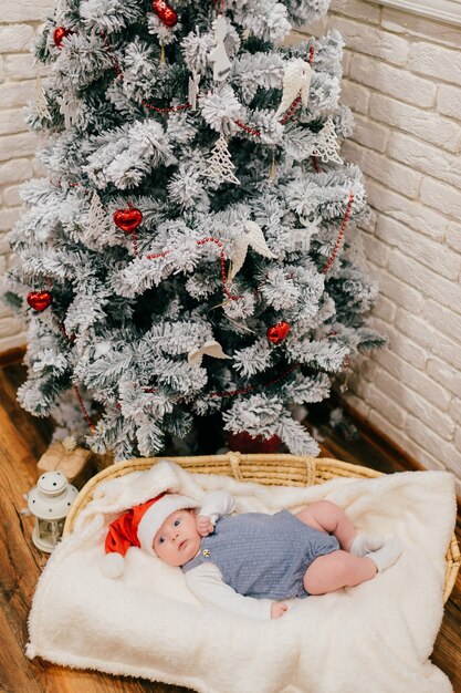 A cute little child in blue pajamas and a red New Year's hat lies in a woven basket near a Christmas tree