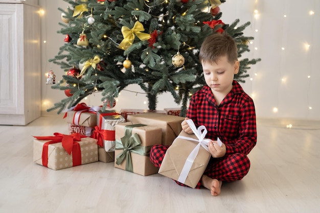 cute little child boy in red pajama with gift boxes sitting under christmas tree at home