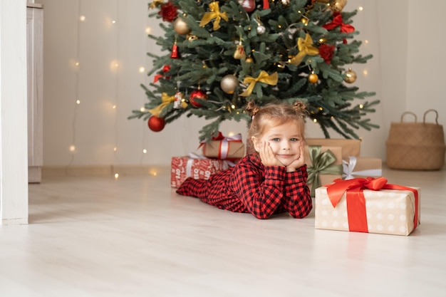 cute little child girl in red pajama lying on floor near christmas tree with gift boxes at home