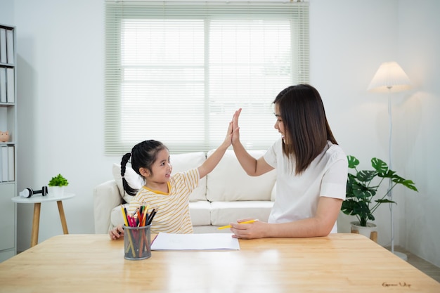 Cute little child and mom hands making give me five while painting with colorful paints Asian girl and mother using crayon drawing color Daughter and mom doing homework coloring cartoon characters