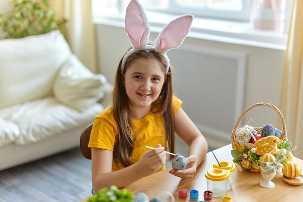 Cute little child wearing bunny ears. Girl sitting sitting at the table in the living room. Kid is painting eggs.