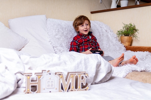 Cute little curly kid boy with smiley face plays games alone on the bed at home stay at home