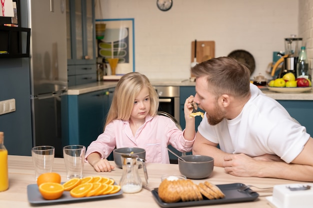 Cute little daughter with long blond hair feeding her father with muesli while both sitting by table and having breakfast