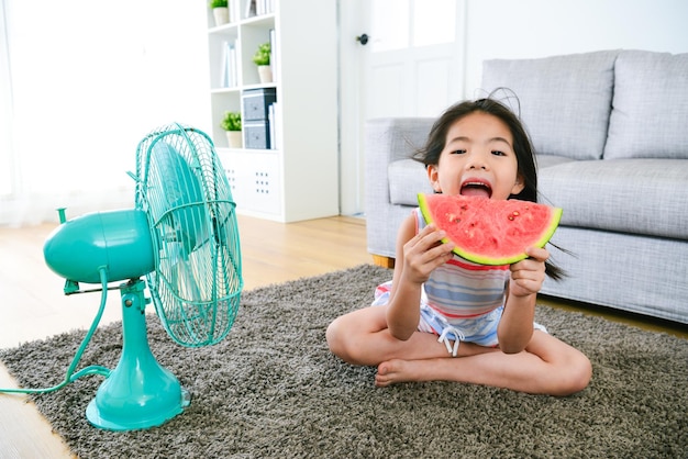 cute little female kid children sitting on floor eating fresh watermelon and blowing electric fan cool wind enjoying summer day.