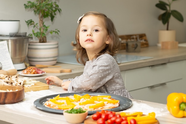 Cute little girl 2-4 in gray dress cooking pizza in kitchen. Kid arranges ingredients on pizza base