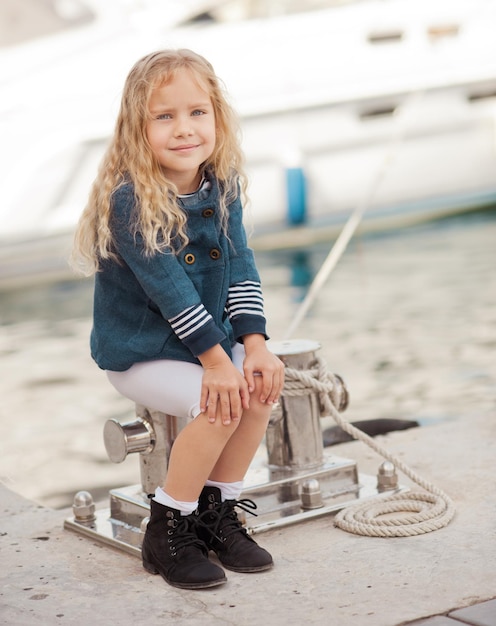 Cute little girl 6-7 year old sitting on seafront outdoors. Smiling child kid. Looking at camera.