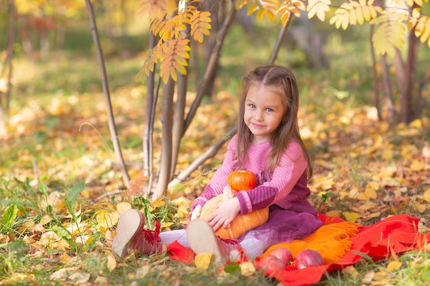 Cute little girl in autumn fall park with orange color leaves and yellow pumpkin